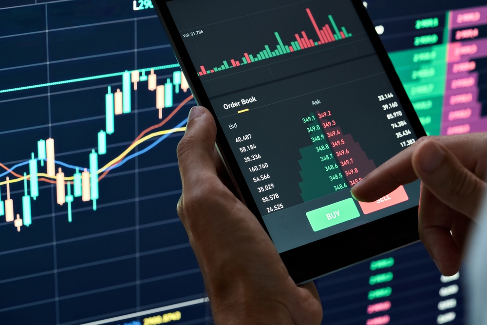 Hands hold a tablet showing a stock trading app with a candlestick chart, order book, and Buy button on screen.