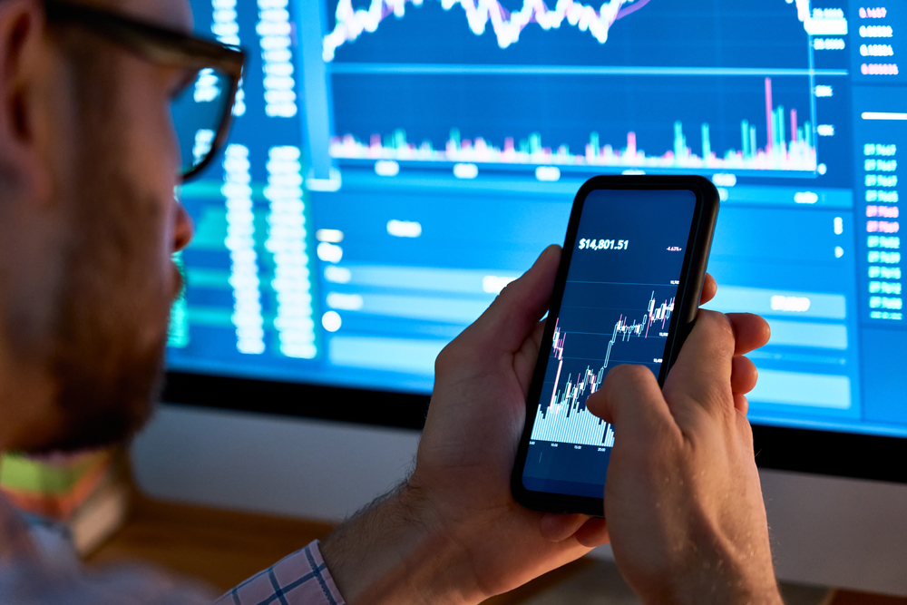 Person holds a smartphone displaying a stock chart while a blue trading dashboard with graphs is visible on a large monitor in the background.